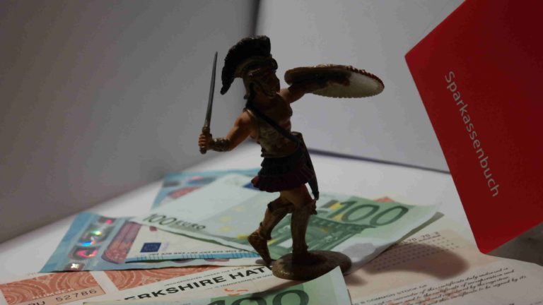 Eine Statur steht auf verschiedenen Geldscheinen und einer Berkshire Hathaway Aktie. Ein Sparkassenbuch steht im Hintergrund.