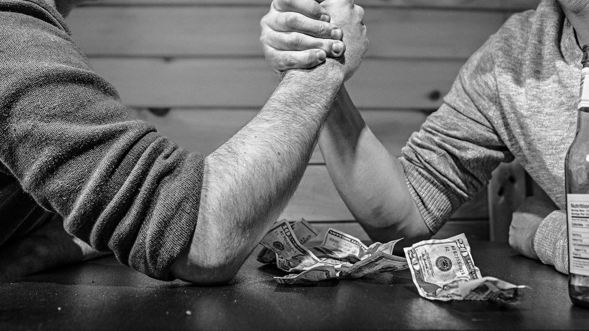 Zwei Männer die beim Armdrücken um Geld kämpfen. Das Geld liegt auf dem Tisch. Nebenher wird Bier getrunken.