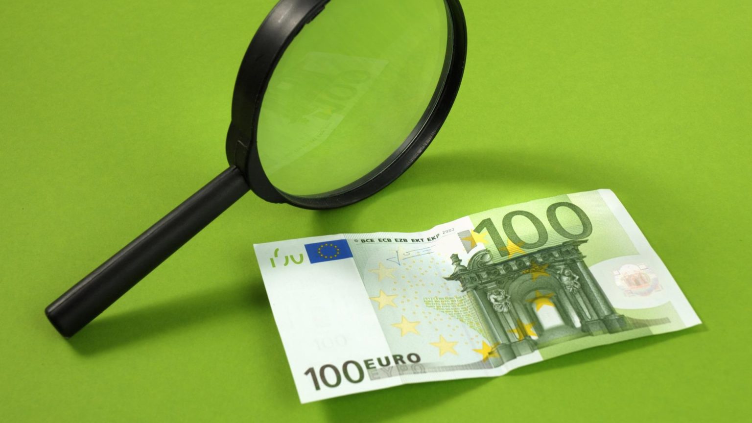 Ein 100€-Schein liegt auf einen grünen Hintergrund und wird von einer Lupe beleuchtet.