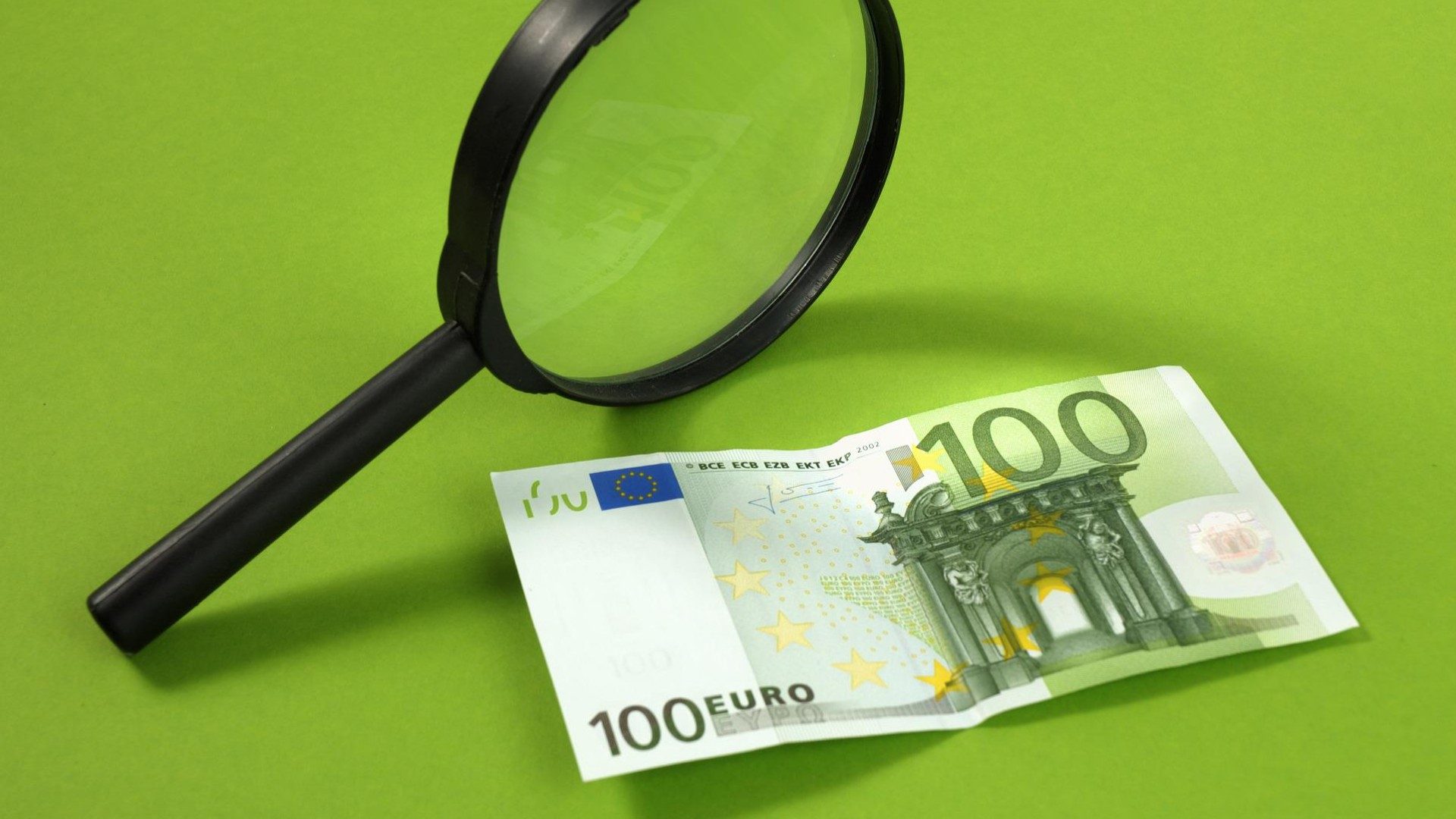 Ein 100€-Schein liegt auf einen grünen Hintergrund und wird von einer Lupe beleuchtet.