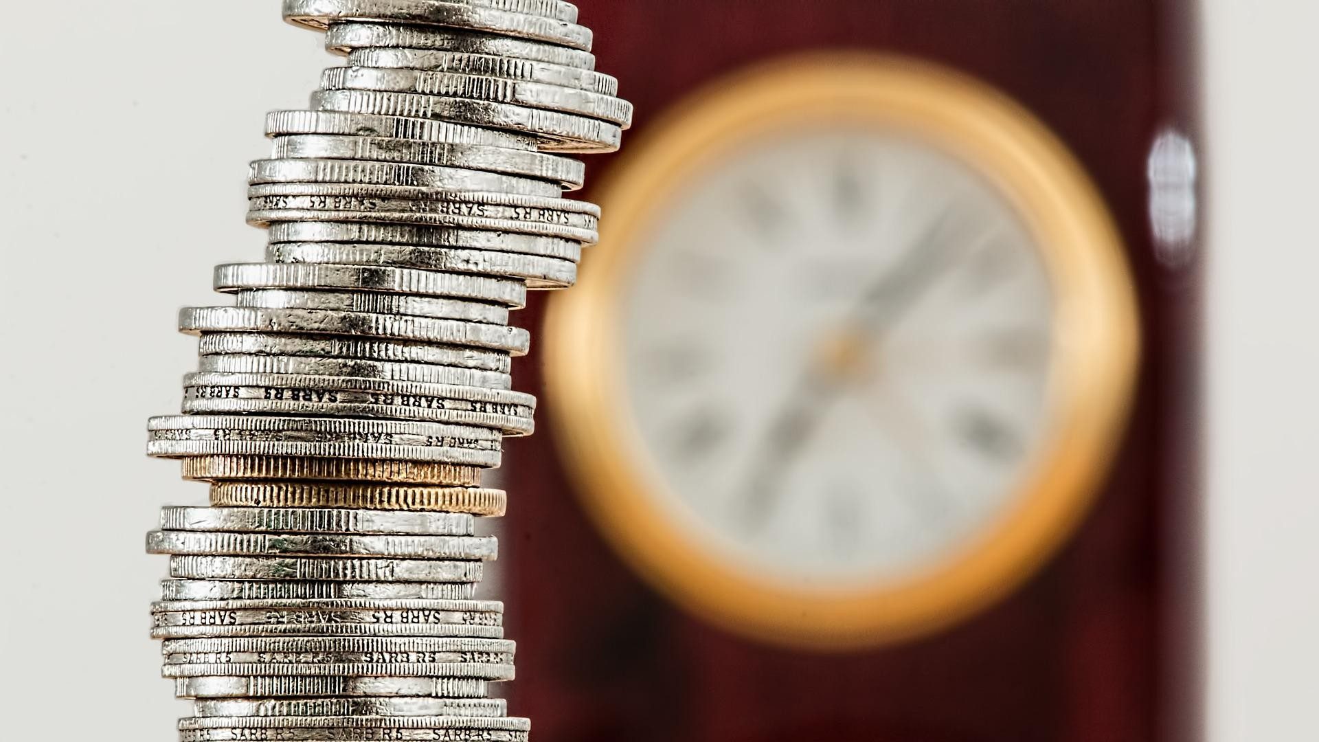 Ein hoher Stapel Geldmünzen vor einer tickenden Uhr repräsentiert Sparen und Sparsamkeit