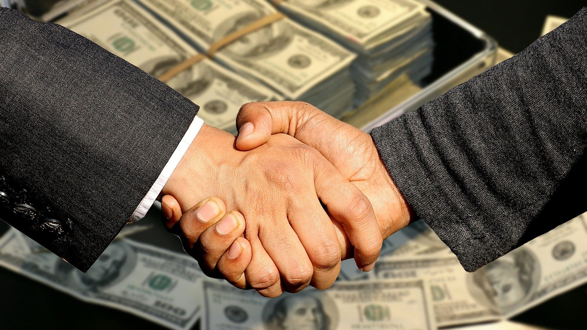 zwei Geschäftsleute geben sich vor einem Stapel Dollar-Geldscheine die Hand zum Deal
