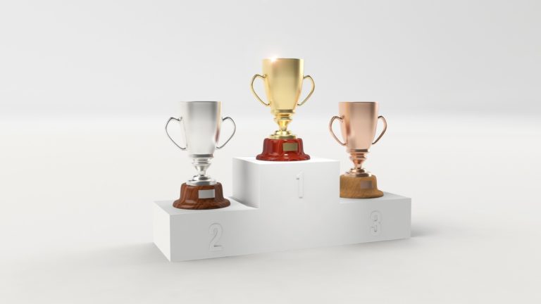 Auf einem Siegerpodest stehen ein Bronze- ein Silber- und ein Goldpokal