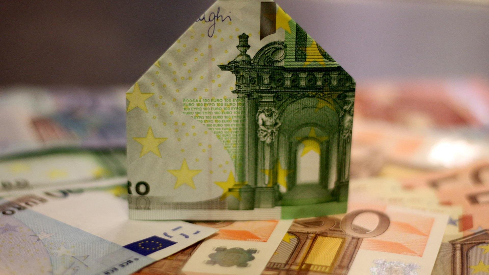 Ein 100€-Schein der zu einem Haus gefaltet wurde steht auf weiteren Scheinen.