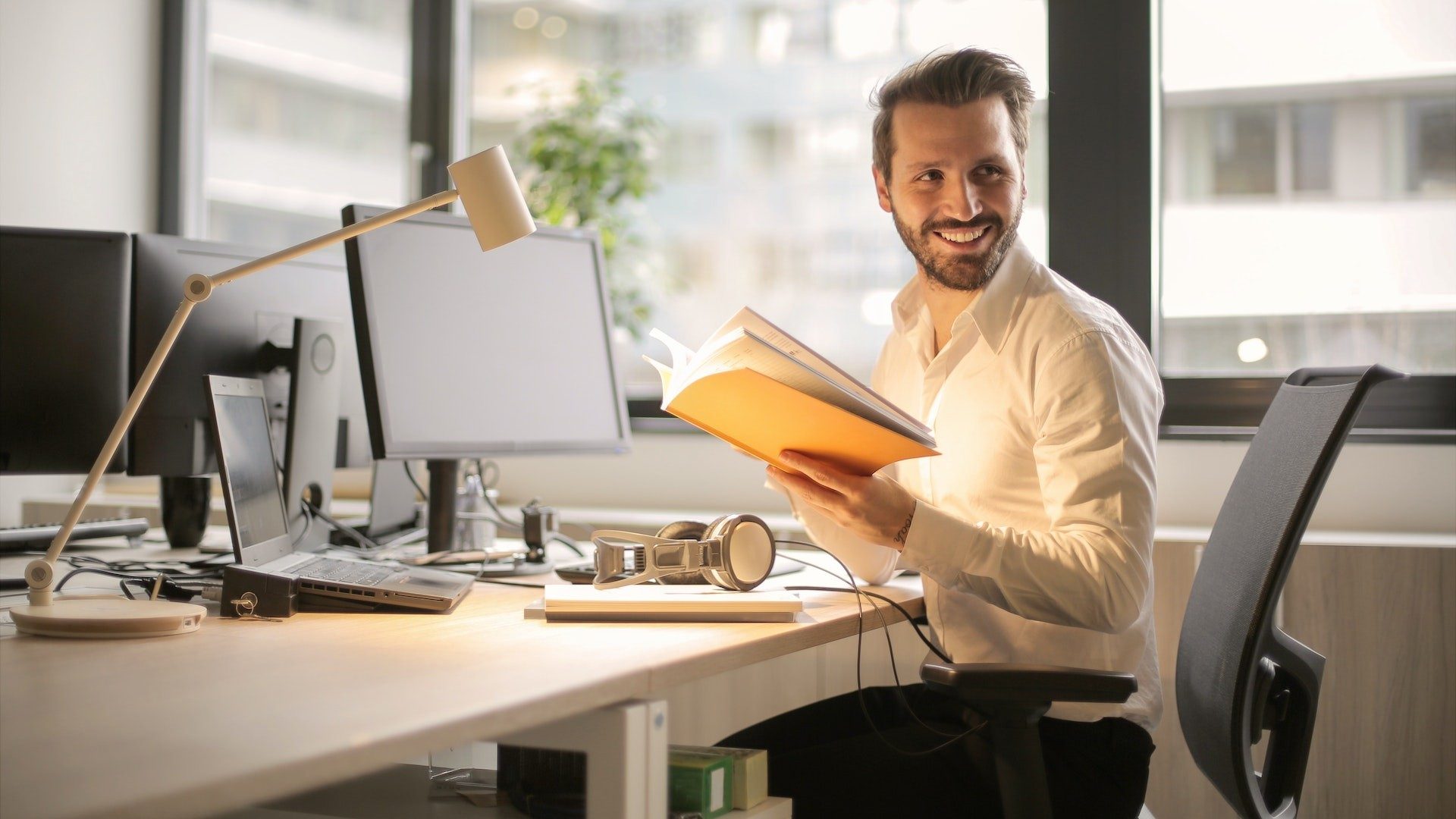 Ein Geschäftsmann am Arbeitsplatz mit Computer hält ein Buch und lächelt freundlich zur Seite