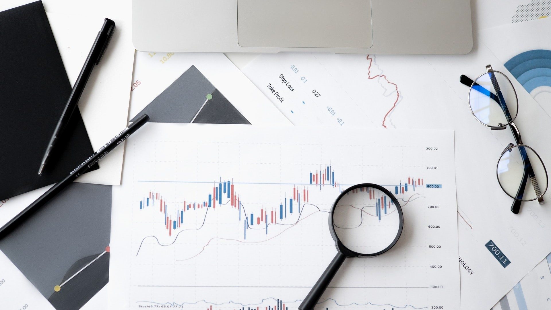 Lupe, Brille und Stifte liegen auf mehreren ausgedruckten Aktien-Charts für die Analyse bereit