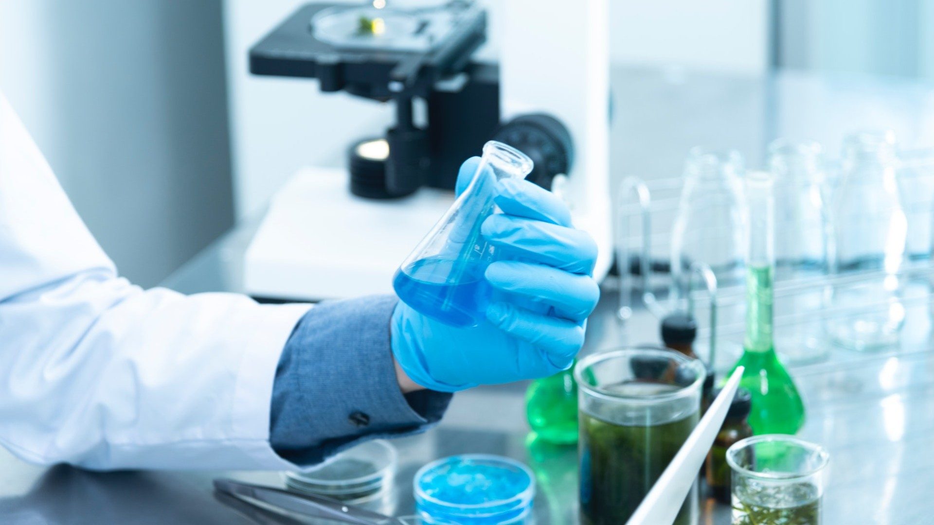 Eine Person im Labor hält eine blaue Reagenz, rundherum weitere Chemikalien und ein Mikroskop