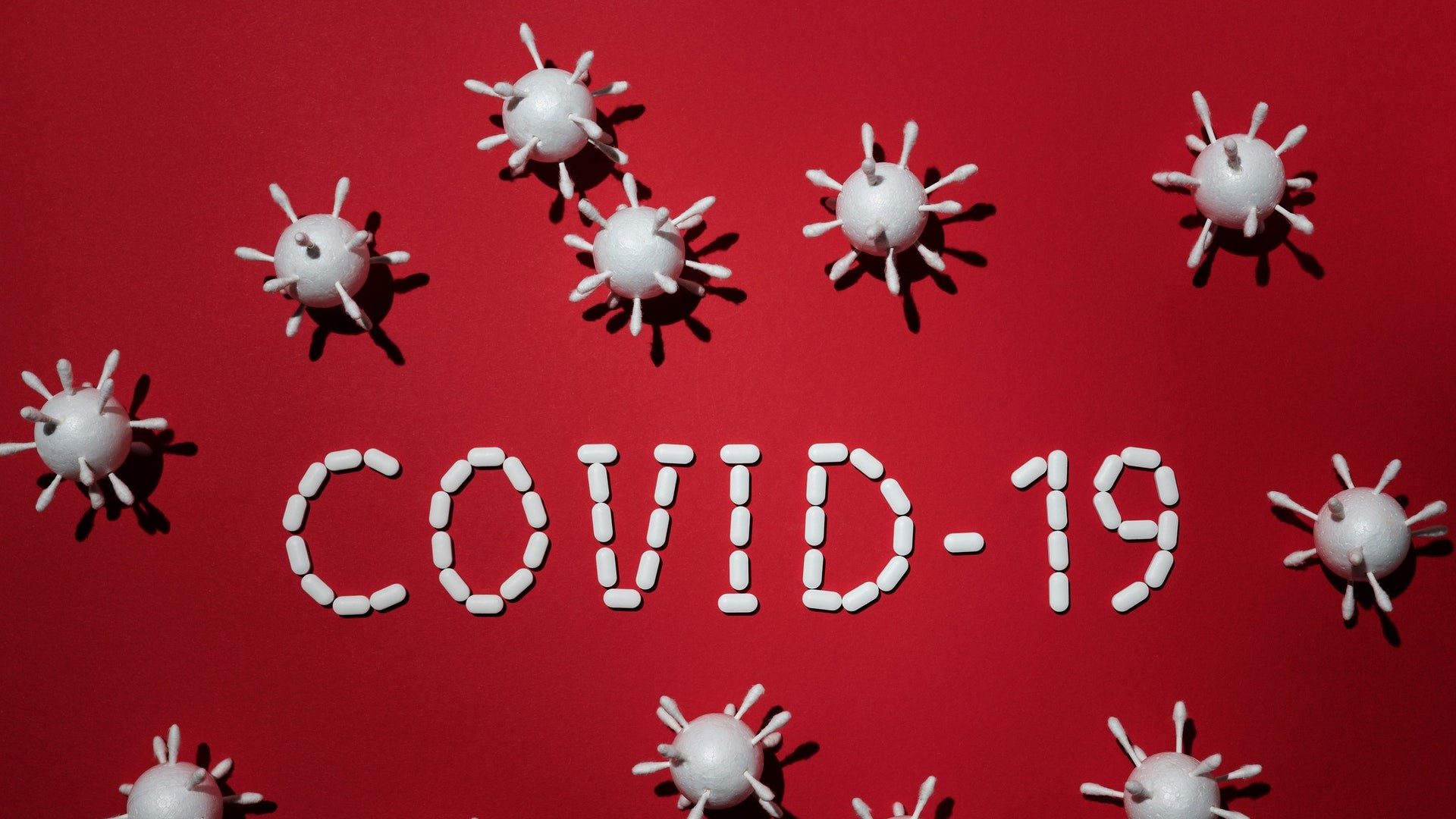 Schriftzug "Covid-19" auf rotem Untergrund mit Pillen und Virus-Figuren