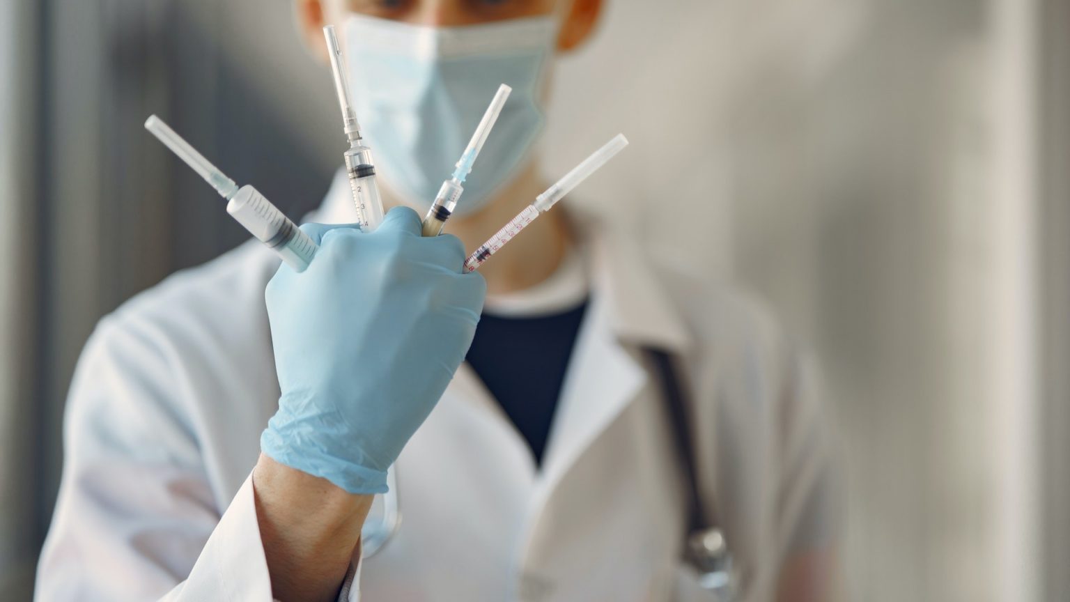 Ein Mann mit Handschuhen und Maske hält vier Spritzen zwischen seinen Fingern