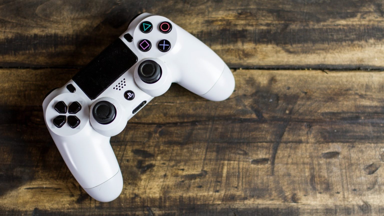 Das Bild zeigt einen weißen Playstationcontroller der auf einen Holztisch liegt.