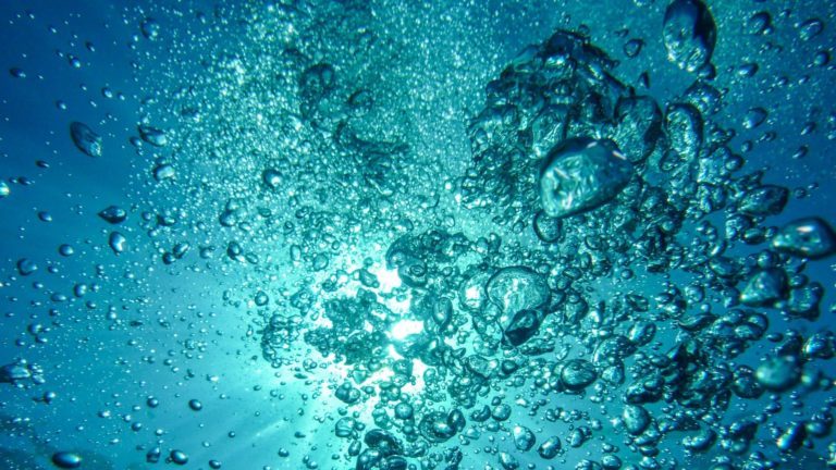 Wasserblasen unter Wasser. Im Hintergrund scheint die Sonne durch das Wasser.