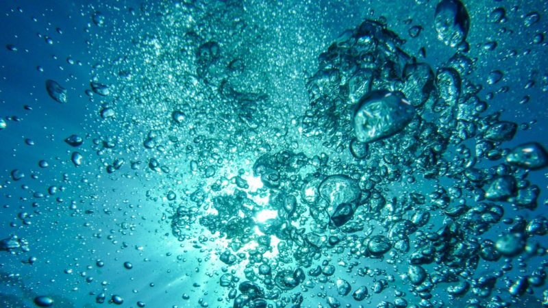 Wasserblasen unter Wasser. Im Hintergrund scheint die Sonne durch das Wasser.