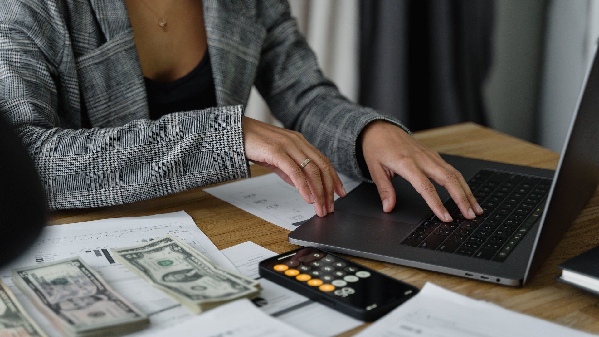 Eine Businessfrau plant ihre Finanzen am Laptop, daneben Rechnungen, Analysen, Taschenrechner und Geldscheine