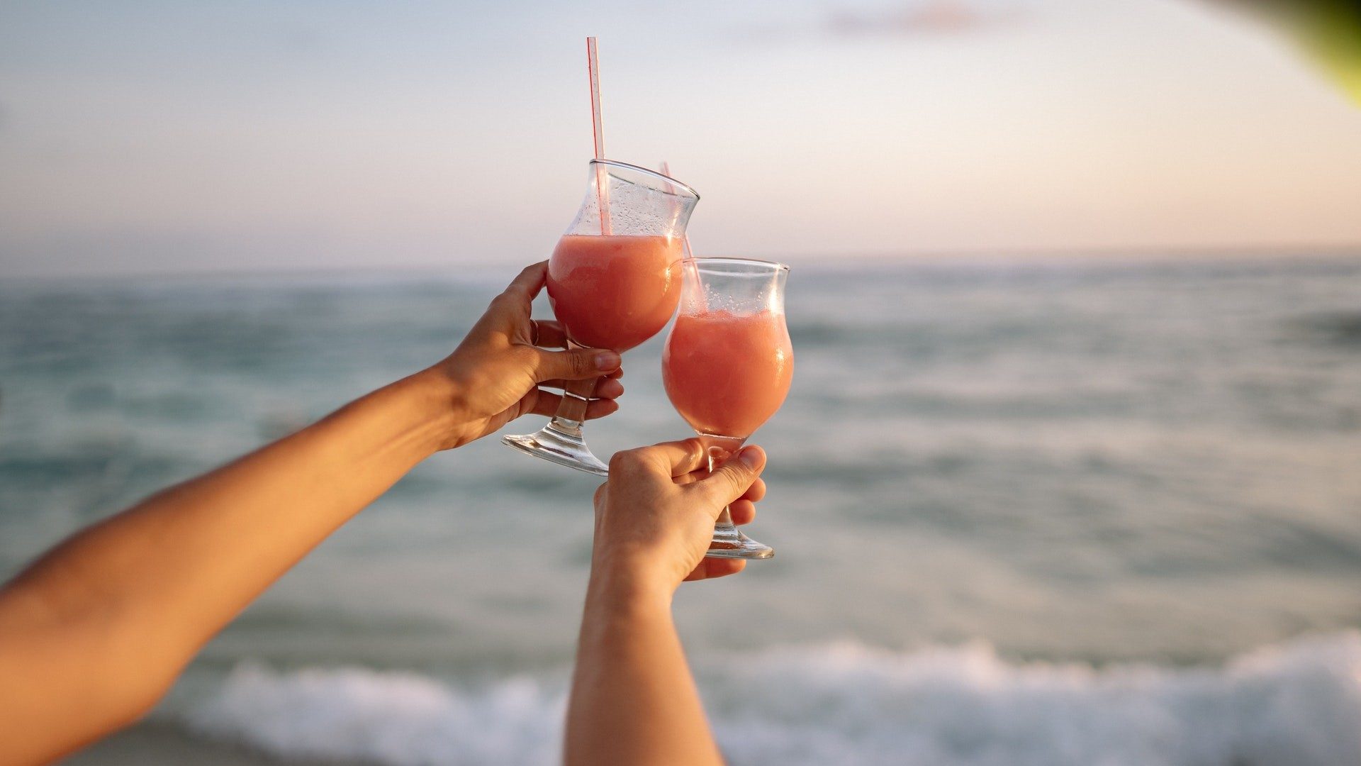 Zwei Personen die einen Cocktail am Strand trinken mit dem Meer im Hintergrund.