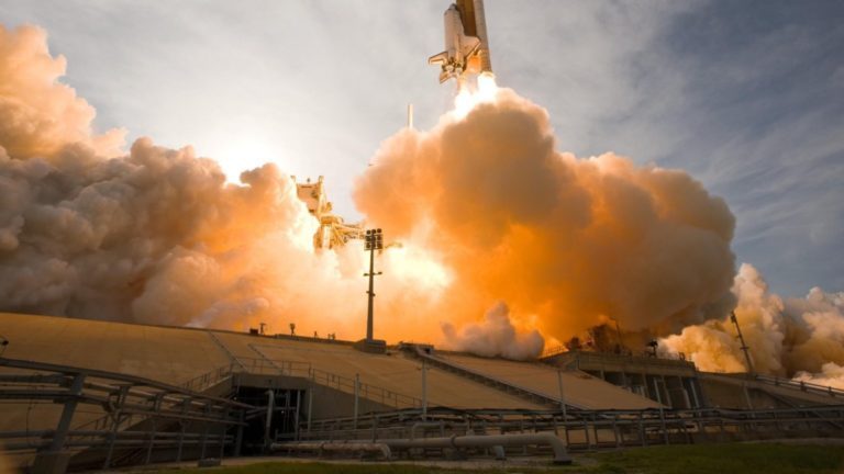 Ein Spaceshuttle startet mit viel Feuer und Qualm von der Erde.