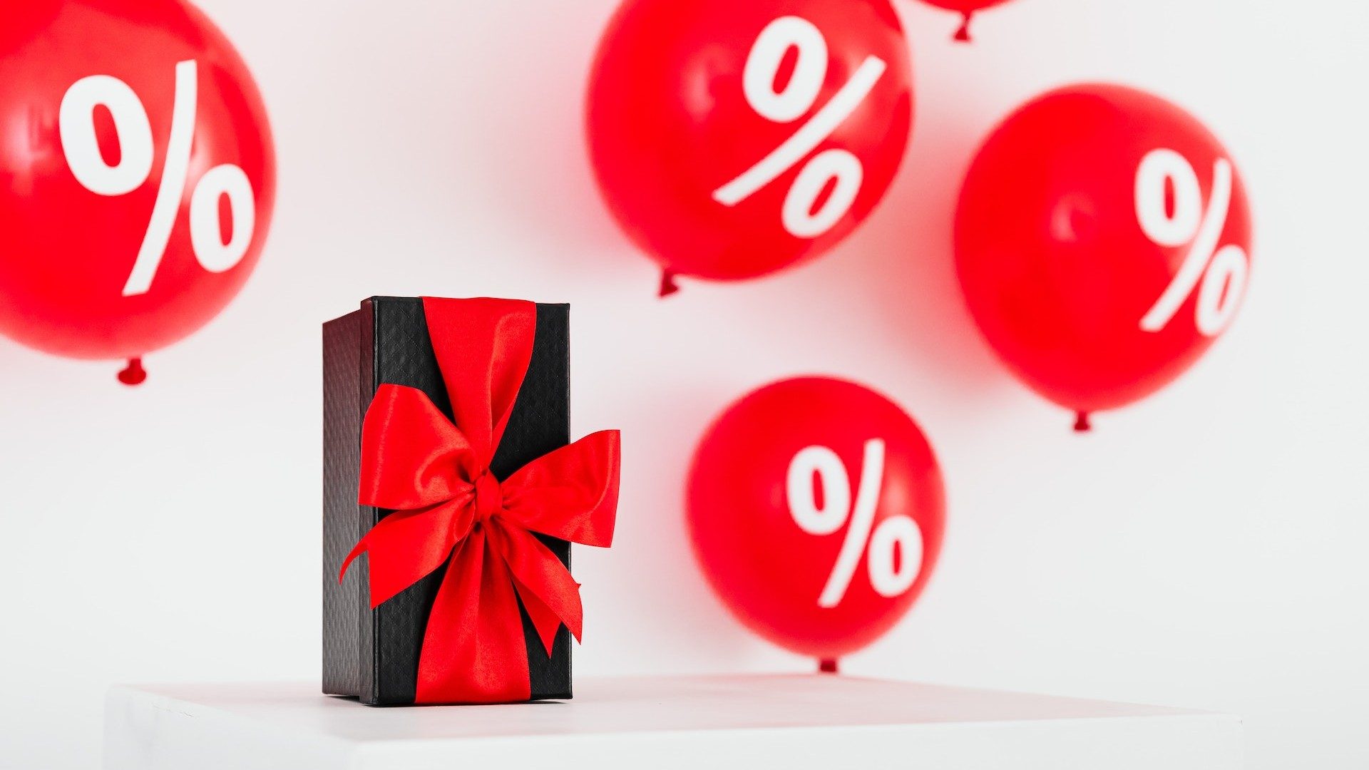 Ein schwarzes Geschenk mit roter Schleife, umgeben von roten Ballons mit Prozent-Zeichen