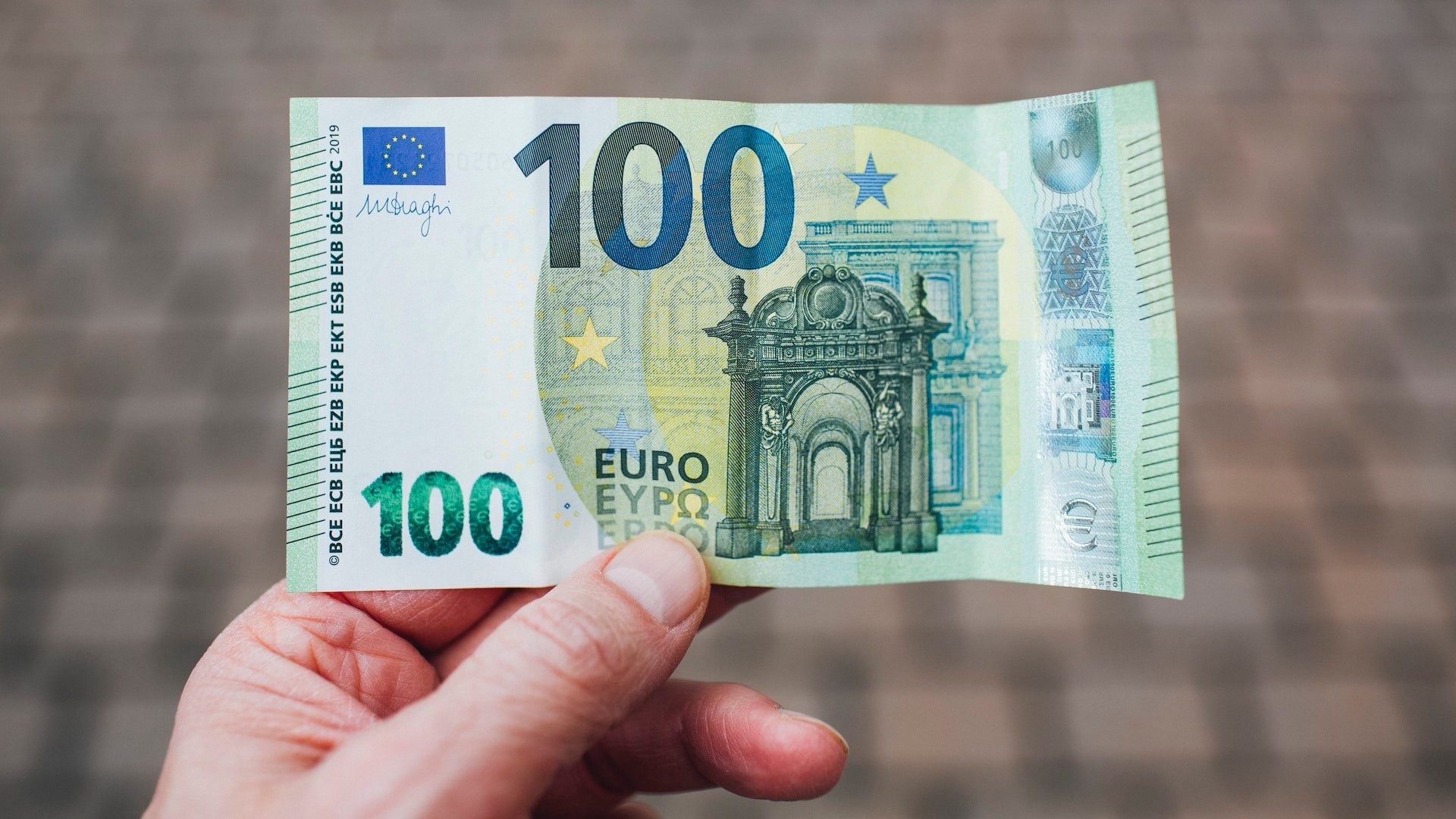 Eine Person hält einen 100-Euro-Geldschein, im Hintergrund Pflastersteine