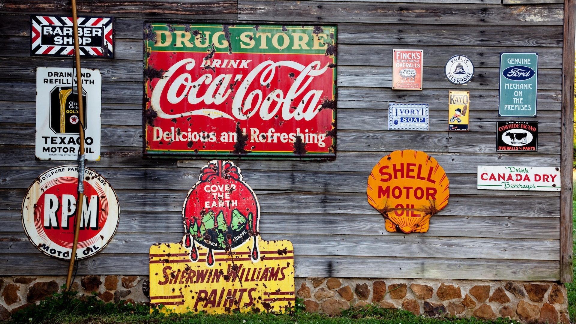 Werbeplaketten von Coca-Cola, Shell, Ford und anderen auf einer grauen Holzwand
