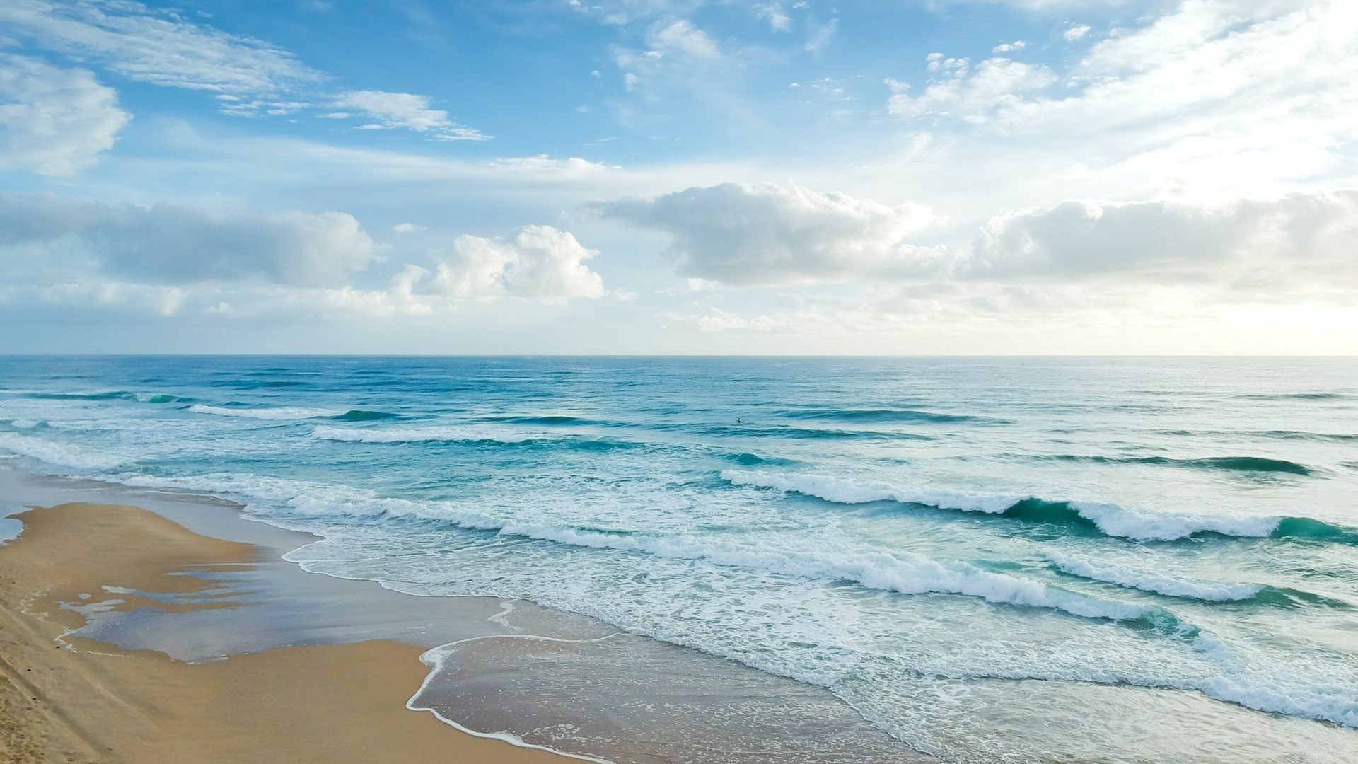 Wellen brechen sich an einem einsamen Strand; im Hintergrund das weite Blau des Ozeans