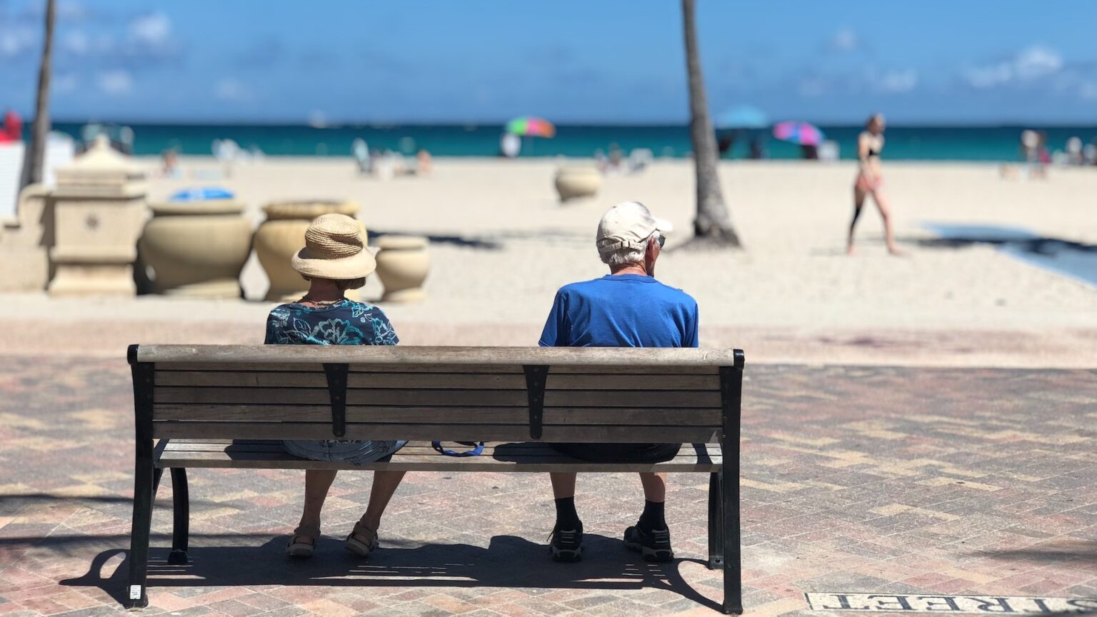 Zwei ältere Menschen auf einer Bank an einer Strandpromenade, im Hintergrund das Meer