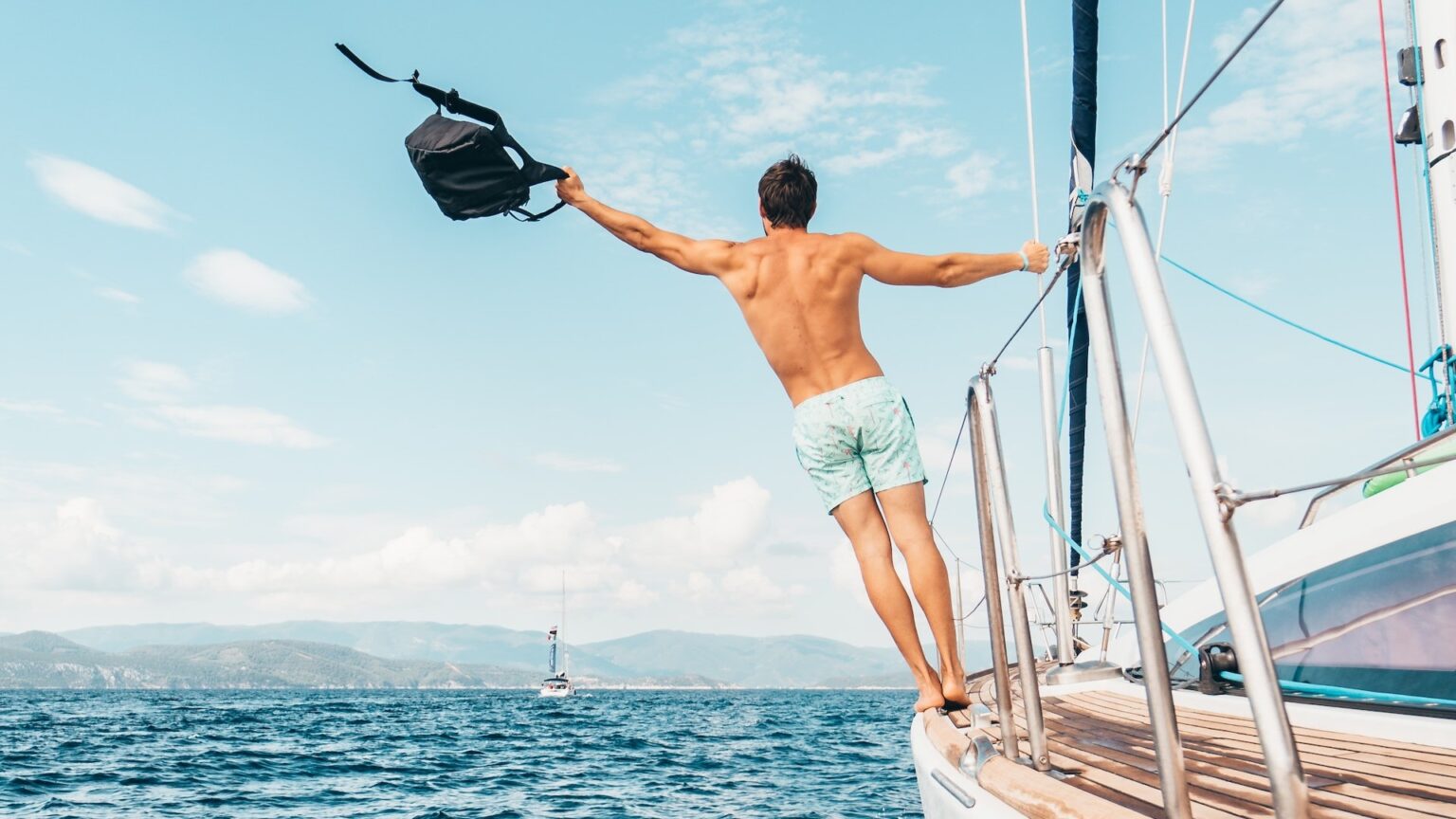 Ein oberkörperfreier Mann lehnt von einer Segelyacht, in einer Hand einen Rucksack