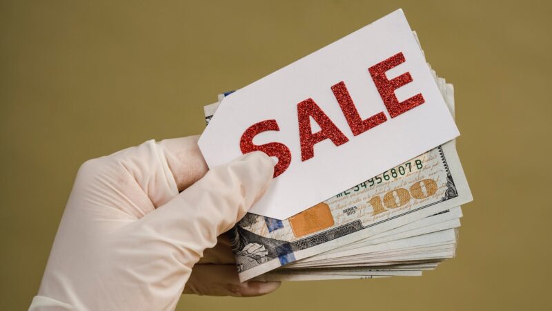 Eine Person hält ein Bündel Geld und ein Etikett mit der Aufschrift "Sale" in der Hand