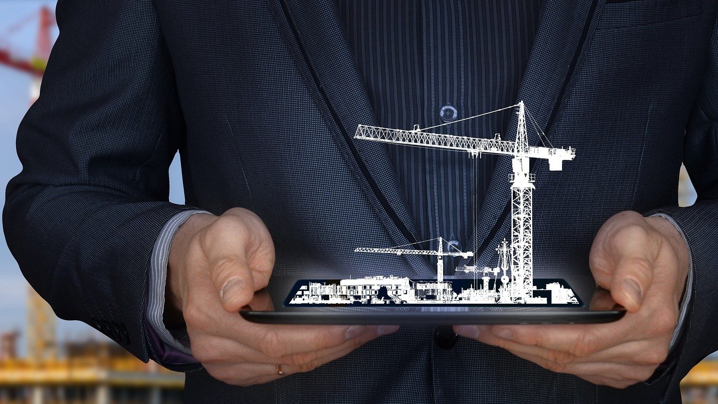 Ein Mann im Anzug hält ein Tablet in der Hand, auf dem illustrierte Kräne und andere Baustellenfahrzeuge stehen