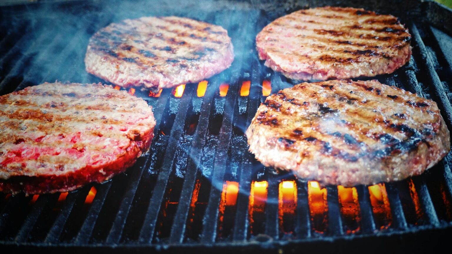 Vier Burger-Pattys werden auf einem Grill gebraten.